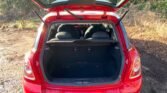 A red MINI with its rear hatch open, revealing a spacious trunk. The back seats are partially folded down, creating more storage space. The vehicle is parked on a dirt road with greenery and trees in the background. Sunlight illuminates the scene, casting shadows on the ground.