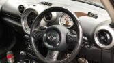 Interior view of a Mini Countryman showing the steering wheel with a Mini logo, dashboard, and center console. The black interior with silver accents contrasts the slightly dirty floor with leaves. A water bottle sits in the cup holder, and through the driver's side window, a brick wall is visible.