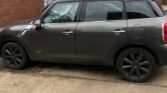 A dark gray Mini Countryman Cooper 2.0 Diesel is parked on a concrete surface near a brick wall. The compact SUV, equipped with automatic transmission and blacked-out wheels, stands in front of a lamppost and a brick building with large windows. A white sign and another car are visible under the cloudy sky.