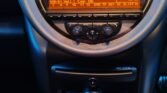 Close-up of a MINI car dashboard featuring a circular speedometer displaying a range from 0 to 160 MPH. Below it, an orange digital screen shows "DAB-D1 Natl | Cap UK." Below the screen are three round climate control knobs and several buttons. Further down, there’s a row of toggle switches and cup holders.