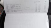 A hand holding an itemized vehicle service invoice for a 2011 Mini One, dated 15/02/2023. The invoice details charges for labor, parts (synthetic oil, water pump, gaskets, etc.), and services totaling £534.92. The manual transmission bill is viewed over a car's steering wheel in an indoor setting.