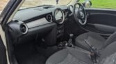 View of the interior of a 2011 Mini One from the driver's side door, which is open. The car, featuring a 1.6 Petrol manual engine, boasts black fabric seats, a dashboard with a circular control panel and display, and a right-side steering wheel. The surroundings include a grassy field and a house.