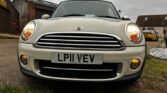 Front view of a white Mini Cooper diesel cabriolet parked outside on a slightly overcast day. The car has a license plate reading "LP11 VEV." The image showcases the distinctive front grille, rounded headlights, and fog lights. Background includes a few buildings and another yellow car on the right.