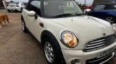 A cream-colored 2011 Mini Cooper cabriolet with a black convertible top is parked on a paved driveway. Its headlights are on. The car has a UK license plate reading "LP11 VEV". Several other vehicles and a golden retriever are visible in the background, along with a house and a cloudy sky.