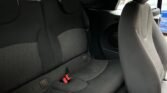 The interior of a 2011 Mini Cooper Diesel Cabriolet is shown, focusing on the rear seats and parts of the front passenger seat. The rear seats are black with a mesh-like center pattern, equipped with headrests and red seatbelt buckles. The passenger seat is also black with gray side padding, partially visible. The car door is slightly open.