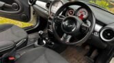 The image shows the interior of a 2011 Mini Cooper cabriolet with black fabric seats, a black leather-covered steering wheel, manual transmission gear shift, and a sleek dashboard with modern controls. The driver’s seat is positioned on the left side, indicating a right-hand drive vehicle.