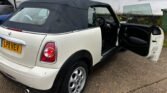 A 2011 Mini Cooper diesel cabriolet with a black soft top is parked on a driveway. The passenger door is open, revealing the interior. There's a green garbage bin and some glass panels near a white building in the background. The car's rear license plate reads "L-P1 VEV.