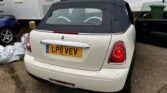 A white MINI Cooper cabriolet with a black soft-top roof is parked on a driveway next to a white building. The vehicle's license plate reads "L P11 VEV." The car's taillights are illuminated. To the left, a portion of another vehicle is visible. There are some items on the ground and green bins nearby.