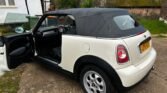 A white 2011 Mini Cooper convertible with a black soft top is parked on a driveway. The driver's door is open, revealing the black interior. The license plate reads "L1P1VEY." In the background, there’s a white house with black trim, green foliage, and an old wooden garage with a moss-covered roof.