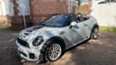 A silver MINI Cooper convertible is parked on a dirt driveway in front of a brick house with white trim. The car's black soft top is down, and it boasts black racing stripes on the hood. The background features a white garage door, part of a house, and trees casting shadows.