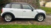 A white MINI Cooper Countryman with a black roof is parked on a concrete driveway. The car features silver alloy wheels and black trim along the bottom and wheel arches. Green grass is in the foreground, and a tall, dry shrub wall stands behind the car. A beige wall and some foliage are in the background.