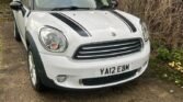 A white MINI Cooper with black racing stripes on the hood and roof is parked on a paved area next to tall brown bushes and greenery. The rear hatch is partially open, revealing the license plate "YA12 EBM." The surrounding area features a mix of fallen leaves and small debris.