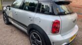 A silver MINI Cooper SUV is parked on a gravel driveway. It has a black roof, tinted windows, and black trim along the sides and rear. The vehicle is positioned at an angle, showing its rear and right side. A brick building and trees are visible in the background.