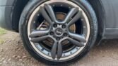 Close-up of a car wheel with a black, multi-spoke rim design. The tire, mounted on a clean alloy wheel with visible bolts around the edge, features the "MINI" logo on the center cap. The surrounding ground is gravelly with some green vegetation peeking through.