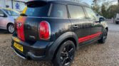 A black MINI Cooper S Countryman with red racing stripes is parked on a gravel surface near a garage. The license plate reads "MJ60 KWD." The garage displays the RAC logo along with signs for sales, service, and parts. Another car and trees are visible in the background.