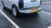 A silver MINI Clubman with black accents is parked in a nearly empty parking lot. The vehicle's UK registration plate reads "FY63 ATV." The parking space is wet with some leaves scattered around, indicating recent rainfall. The background shows a few buildings and faintly overcast skies.