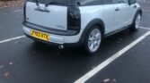 A silver MINI Cooper with a UK registration plate "FY63 ATV" is parked in a parking lot. The car has black-tinted windows and tail lights, positioned in a marked space. The overcast sky and wet asphalt suggest recent rain, with scattered leaves on the ground.