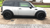 A side view of a white MINI Cooper car with a black roof and black wheels parked on a brick-paved driveway. The background shows suburban houses with brick facades, green lawns, and a tree on the left. The car's windows are tinted, and the driveway appears slightly wet.