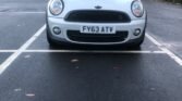 A silver MINI Cooper is parked in an outdoor parking lot. The car has a black roof and side mirrors, and its front license plate reads "FY63 ATV." The parking lot is mostly empty with scattered fallen leaves on the ground. In the background, there are bare trees and a building.