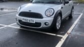 A silver MINI Cooper with the license plate "FY63 ATV" is parked in a near-empty lot. The car has a black roof, and the sky in the background appears overcast, suggesting a typical cloudy day. The ground is wet, indicating recent rain, and trees are visible in the distance.