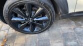 Close-up of a black wheel with a glossy finish on a white 2013 MINI Hatchback, 1.6 Cooper Petrol Manual, parked on a paved surface with gray stones. The tire displays the brand "QDZAIWT" and the code "DX640." A section of the wheel well is also visible.