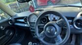 The interior of a 2013 MINI Hatchback features a black dashboard and leather seats. The steering wheel displays the Mini emblem, and the dashboard includes two large dials for the speedometer and tachometer. A pink air freshener hangs from the rearview mirror, with a red car visible through the windshield.