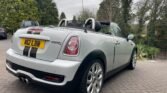 A grey convertible MINI Cooper S with a black soft top parked on a paved driveway. The car has a dual exhaust, rear stripes, and a UK registration plate reading "MK12 LBB." The setting features lush greenery and tall trees in the background, with other vehicles visible nearby.