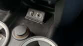 Close-up of a MINI's center console. Two cup holders with Union Jack designs are visible at the bottom. Above the cup holders are two circular controls, likely for climate settings. Above the controls is a power socket and a toggle switch panel with various buttons and knobs.