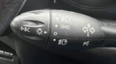 Close-up of a MINI's multifunction steering column stalk, positioned left of the steering wheel. The stalk features various symbols for controlling turn signals, lights (headlights, parking lights), and a control labeled "BC." Part of an air vent and the steering wheel cover are also visible.