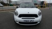 A white MINI Cooper with the license plate YN63 JPJ is parked in a mostly empty parking lot. The background features a large, modern, industrial building with a flat roof and metal exterior. The sky is overcast, and several yellow bollards and other parked cars are visible.
