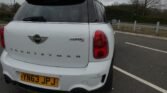 A white MINI Cooper S Countryman is parked in a nearly empty parking lot. The vehicle's rear end is shown, displaying its model badges and a UK license plate reading "YN63 JPJ." The sky is cloudy, and there are bare trees and a few light poles in the background.