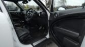 Interior view of a white MINI, showing the driver's side door open. The seats are black leather with matching interior accents. The dashboard features a large central display and round air vents. The floor is carpeted, and the steering wheel and controls are visible. Outside, trees are visible.