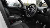 The interior of a compact MINI with black seats and dashboard is shown. The steering wheel and gear shift are visible. Various controls and a smartphone holder are attached to the dashboard. The outside view shows other parked cars and trees, hinting at an overcast day.