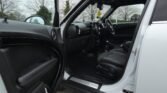 Interior view of a MINI with the driver-side door open, showcasing the black leather seats and dashboard. The door panel has built-in controls. A tree-shaped air freshener hangs from the rearview mirror. The exterior is white, and the background shows a cloudy day with trees.