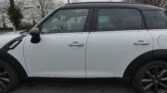 A white MINI Cooper with a black roof and black trim sits parked on a road. The car's windows are slightly tinted, and its black alloy wheels are visible. Trees without leaves stand in the background, with some buildings partially visible behind them under an overcast sky.