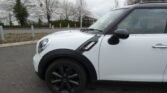 Side view of the front section of a white MINI Cooper S in a parking lot. The car boasts black accents, including the side mirrors, wheel arches, and wheels. Trees and a large white building are visible in the background, along with metal bollards and a concrete border.