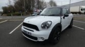 A white MINI Cooper with black trim and black wheels is parked in an empty parking lot. The vehicle has UK plates reading "YN63 JPJ." There are trees and a FedEx station in the background on a cloudy day. A truck is visible on the right.