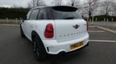 A white MINI Cooper Countryman with a black roof and tinted windows is parked in a nearly empty outdoor parking lot. The car's license plate reads "YN63 JPJ." The sky is overcast, and bare trees and a hedge line are visible in the background. A yield sign is also visible.