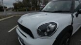 A white MINI Countryman is parked in a lot with its front right headlight prominently visible. The sky is overcast, and leafless trees are in the background. A FedEx truck is partially visible, driving on the road behind the car. Road signs and poles are seen to the left.