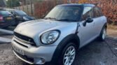 A silver MINI COUNTRYMAN COOPER SD is parked on a wet driveway in front of a tall hedge with autumn foliage. The car, with UK license plate VF64 VLS, has a black roof and mirrors, capturing its clean, shiny exterior in daylight. In the background, there's a wooden fence and other vehicles.