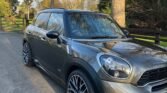 A sleek, gray MINI Cooper is parked on a tree-lined street. The car features sporty black alloy wheels with red brake calipers and a modern design with tinted windows. The surrounding area is serene with tall trees and a wooden fence in the background, suggesting a rural or suburban setting.