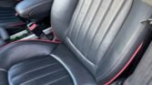 Close-up view of the interior of a MINI, focusing on two black leather seats with red stitching. The front seats feature a combination of smooth and textured leather, with horizontal stitching patterns on the seating surfaces. The armrest and seat adjustments are visible between the seats.