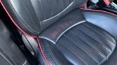 Close-up of a MINI's leather seat, showcasing black upholstery with red piping along the edges. The seat features vertical stitching and appears to be well-padded. Part of the car's interior, including the center console and seat adjustment controls, is also visible.