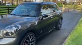 A metallic gray MINI Cooper SUV is parked on a paved driveway bordered by a green lawn. The vehicle has sporty black and silver wheels with red brake calipers. In the background are large, leafless trees and a wooden gate, indicating a winter or early spring setting in a residential area.