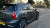 A gray MINI Countryman with UK license plate "LM14 UHH" is parked on a road with bare trees and a clear sky in the background. The MINI has distinctive black and silver alloy wheels, and the shadow cast by the car and trees indicates the sun is low in the sky.