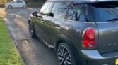 A sleek, gray MINI Countryman is parked on a suburban street near a stone house surrounded by trees. The sun is low in the sky, casting long shadows. In the background, a white car is parked on the opposite side of the street. The road is clear, and the grass is lush and green.