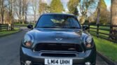 A dark gray MINI Cooper S with a license plate reading "LM14 UHH" is parked on a paved road flanked by grass and wooden fences. The background includes deciduous trees with sparse leaves, evergreens, and a blue sky with scattered clouds. Shadows suggest late afternoon sunlight.