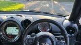 Interior view of a MINI Cooper car showcasing the steering wheel with the iconic MINI logo, a speedometer, and a dashboard featuring a navigation screen displaying a map. Sunlight filters through the windshield, casting light on control knobs. Outside, a blue van is parked beside lush trees and greenery.