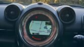 The image shows the dashboard of a MINI car with a digital navigation screen displaying a map at the center. The dashboard features two symmetrical air vents on either side of the screen. The car is driving on a road with trees and the setting sun visible through the windshield.