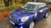 A parked blue MINI Cooper with a white roof is shown on a wet street. The car is surrounded by autumn trees with fallen yellow and brown leaves scattered on the road. The car's license plate reads "M104 JLO".